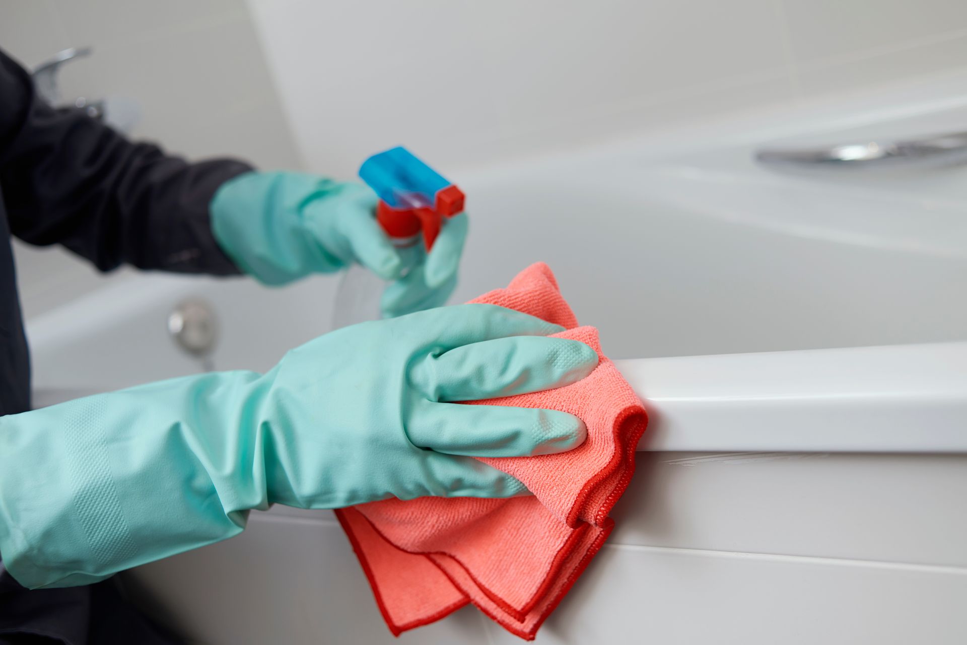 Gloved hands cleaning a white bathtub with a spray bottle and red cloth