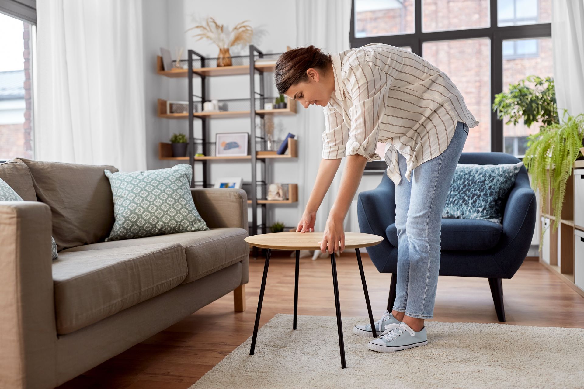 Person adjusting a small table in a bright living room with a gray sofa and blue chair