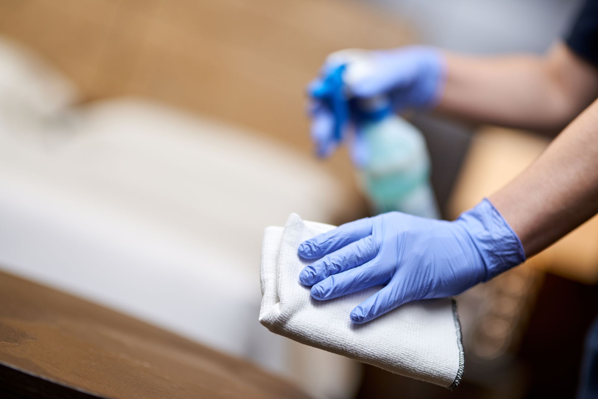 Gloved hands wiping a wooden surface with a white cloth and spray cleaner