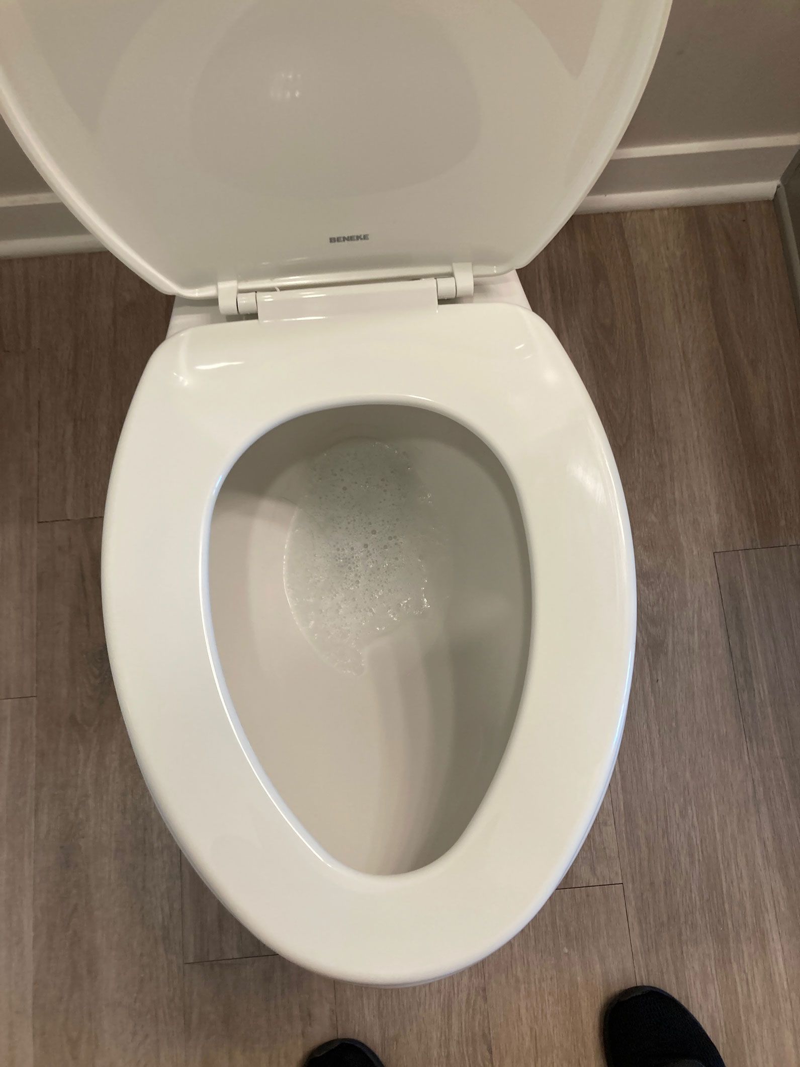 Open toilet in a bathroom with white porcelain seat and lid, brown wood floor, and a black shoe visible.