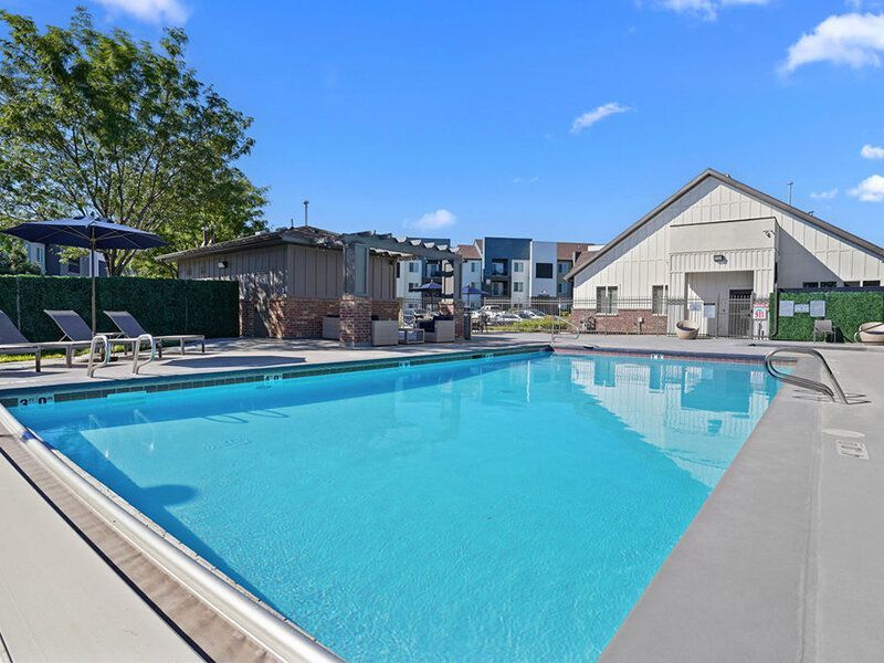Pool with lounge chairs on a sunny day at Wilshire Place, featuring apartments in West Jordan, UT. A building and trees are in the background.