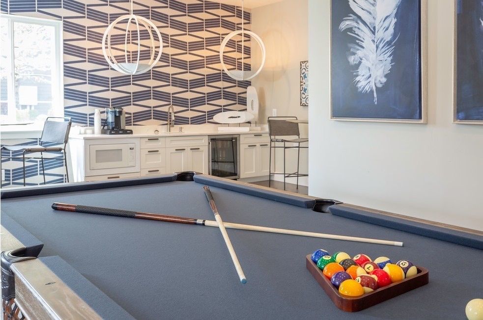 Indoor community game room with a pool table, cues, and a kitchenette in the background at Wilshire Place, featuring townhomes for rent in West Jordan, UT.
