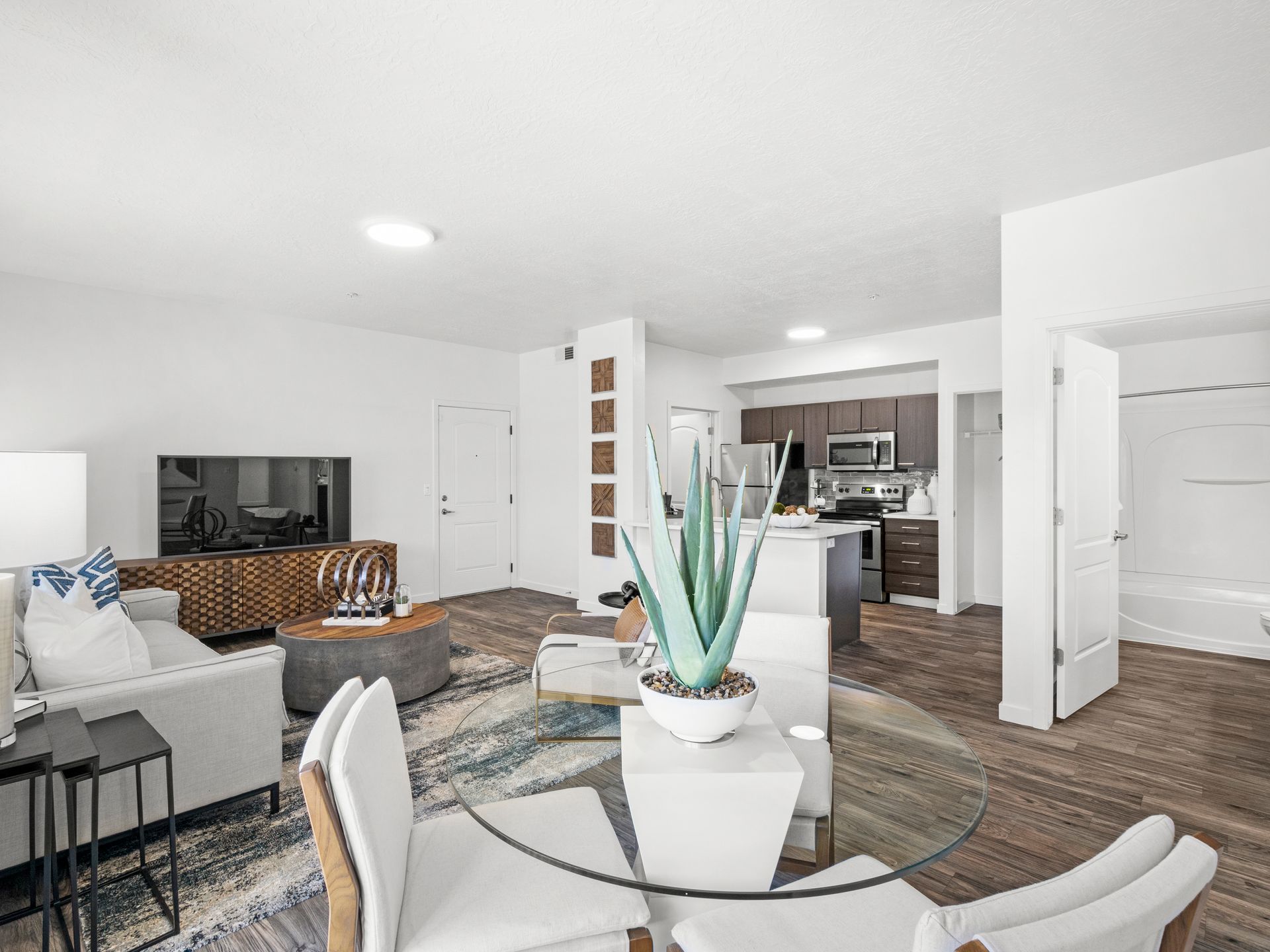 Open-concept living room with sofa, TV stand, round glass dining table, and visible kitchen at Wilshire Place, featuring townhomes near Salt Lake City in West Jordan, UT.