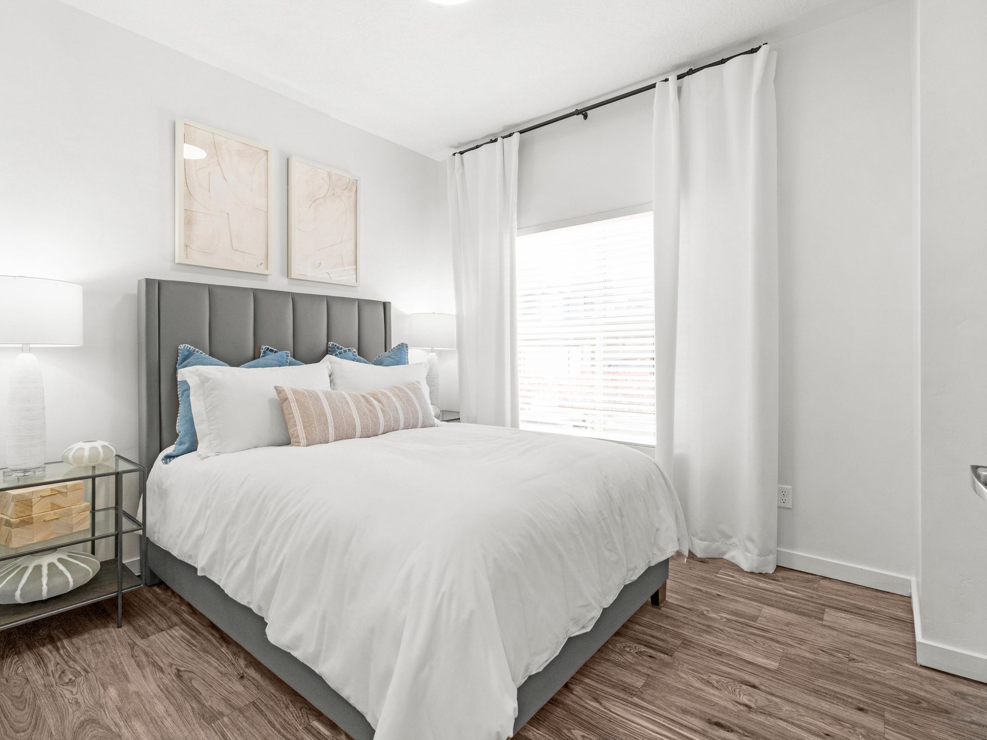 Bright bedroom in a modern apartment with a gray upholstered bed, white bedding, and white curtains at Wilshire Place, featuring apartments near Salt Lake City in West Jordan, UT.