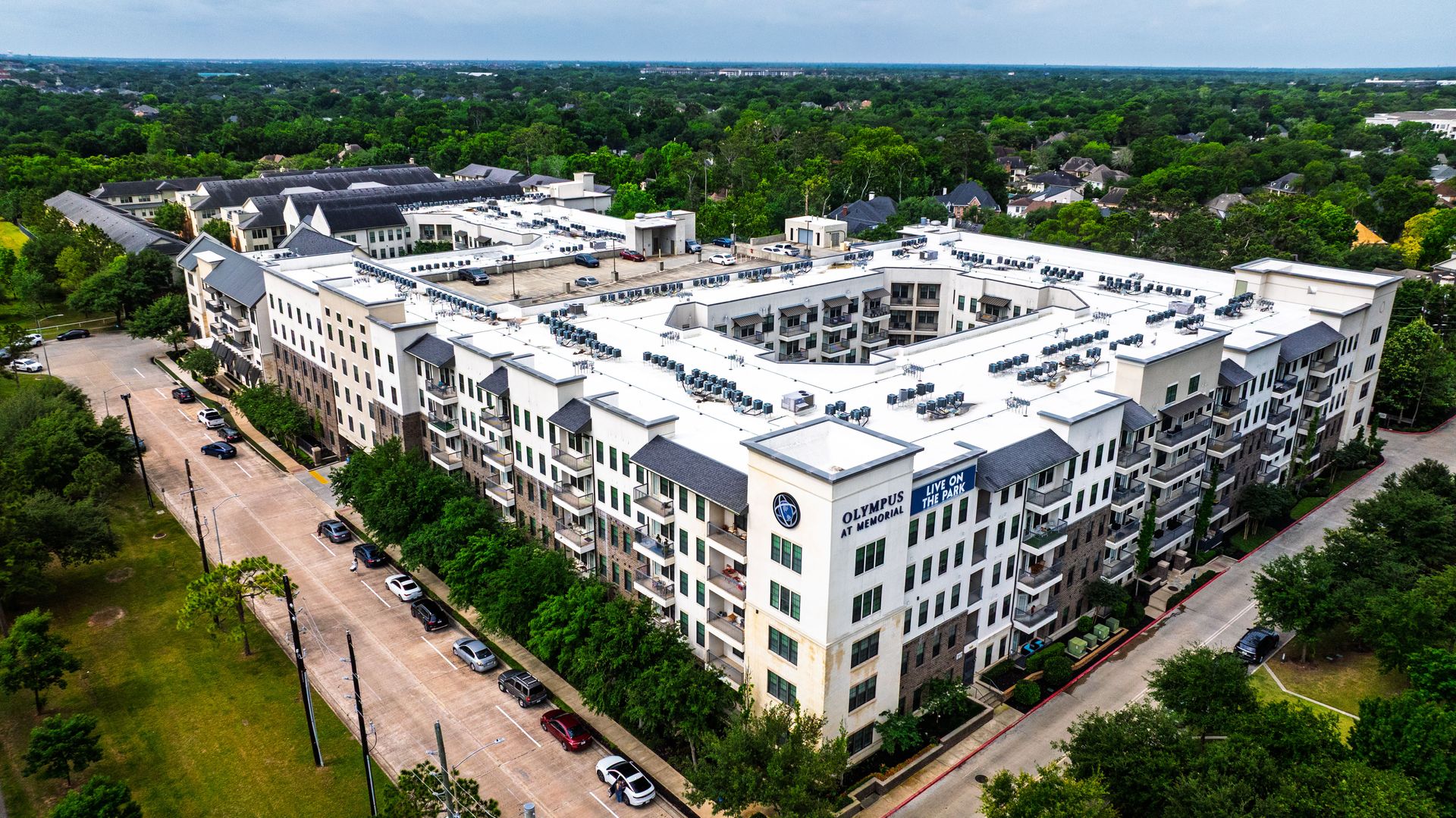 Aerial view of a large white apartment complex surrounded by trees and roads at Olympus at Memorial, offering apartments in West Houston, TX.