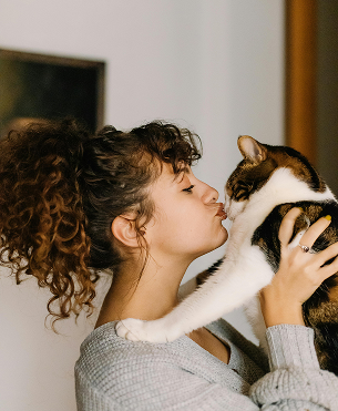 A person with curly hair in a high ponytail wearing a gray sweater leans in to kiss a white and brown tabby cat.