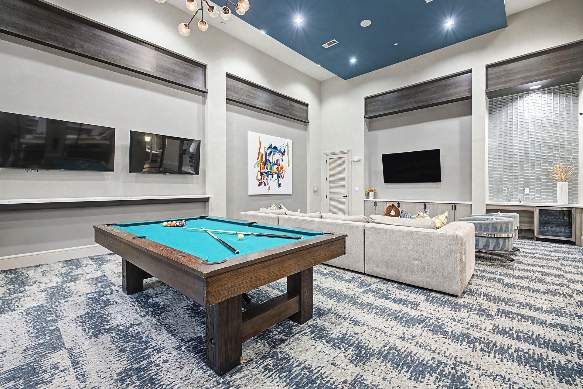 Modern game room with pool table, sofas, wall TVs, and blue patterned carpet at Olympus at Memorial, offering townhomes for rent in the Memorial area of Houston, TX.