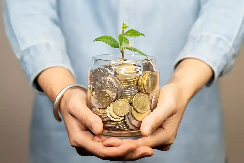 A person is holding a jar of coins with a plant growing out of it.