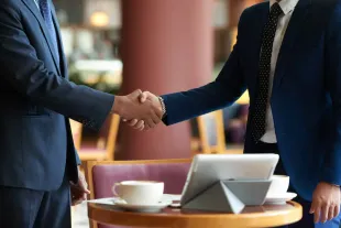Two businesspeople shaking hands across a table with a laptop and coffee cup