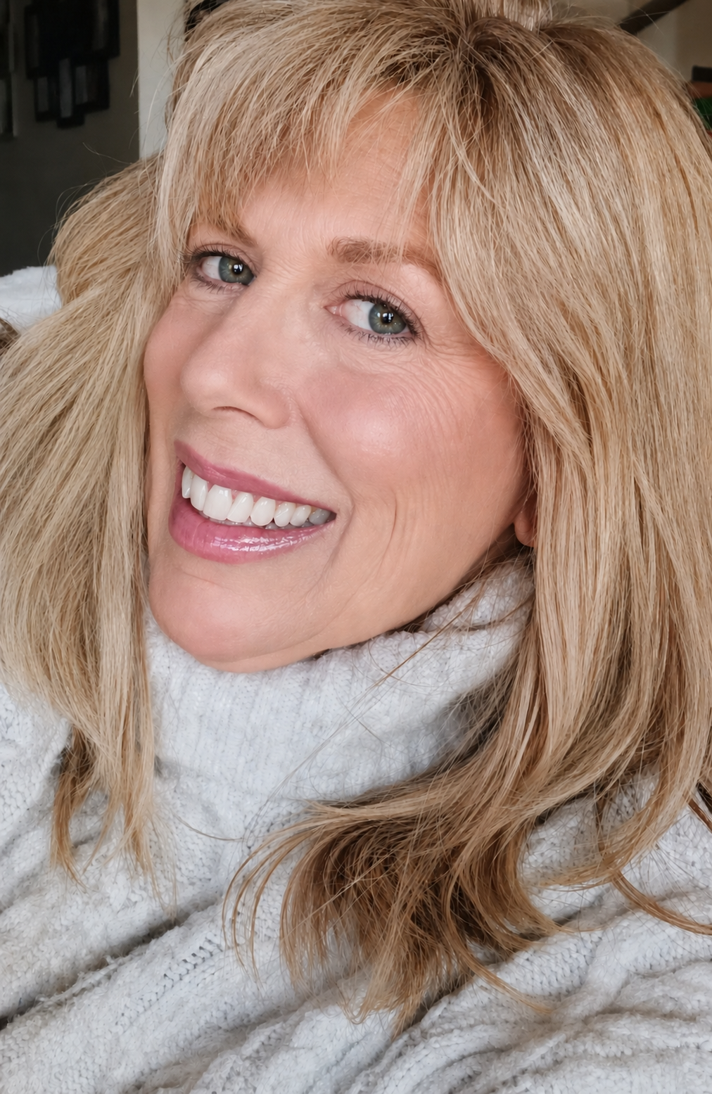 Smiling woman in a white sweater with blonde hair, posing indoors against a dark background