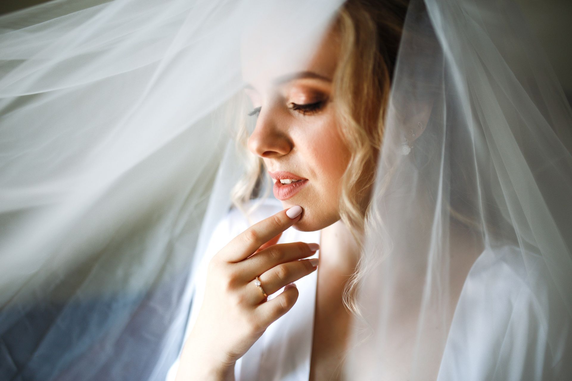 Bride in a white veil, softly lit, touching her chin in a close-up portrait