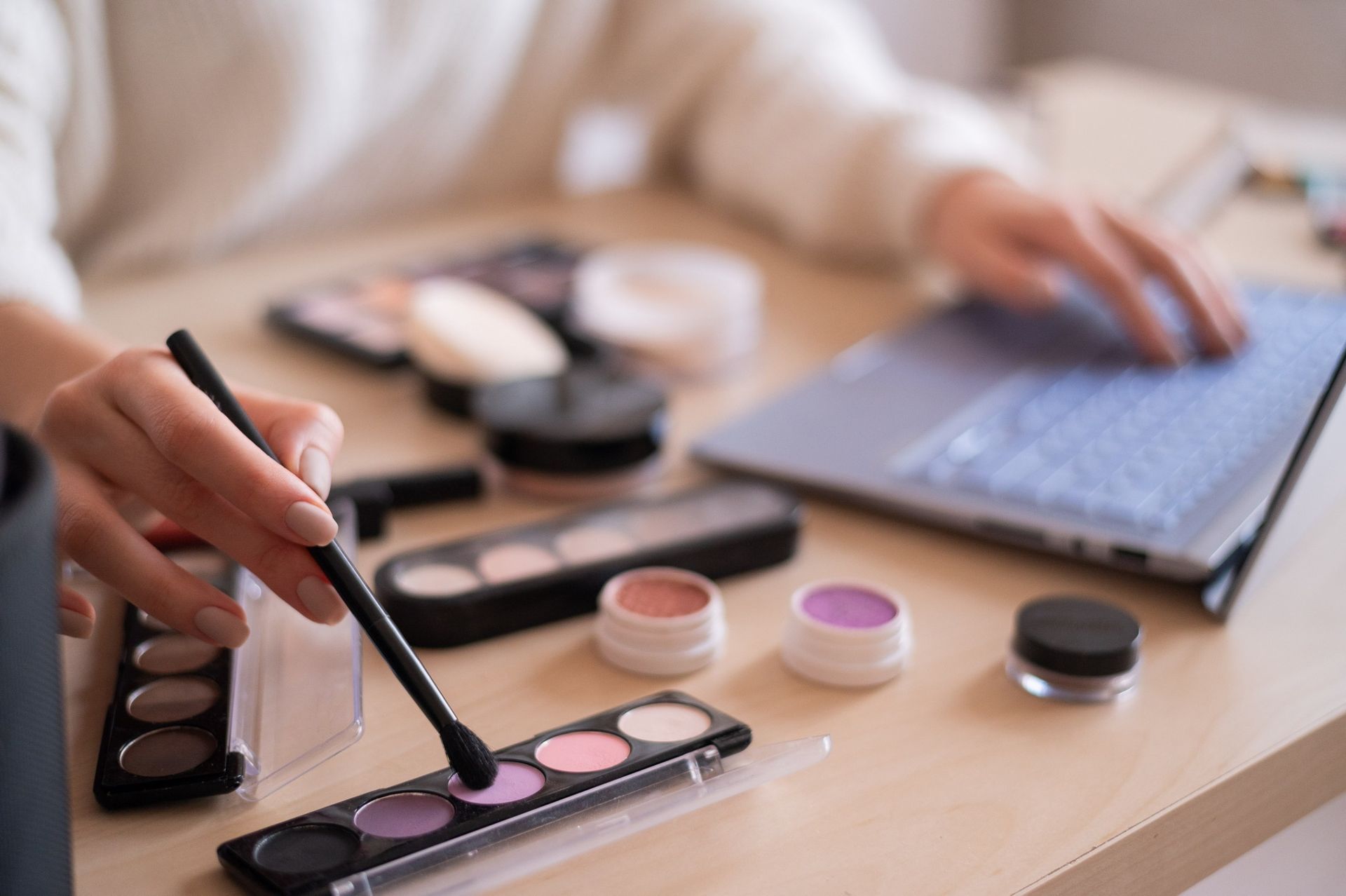 Hands using a makeup palette and brush beside a laptop on a desk