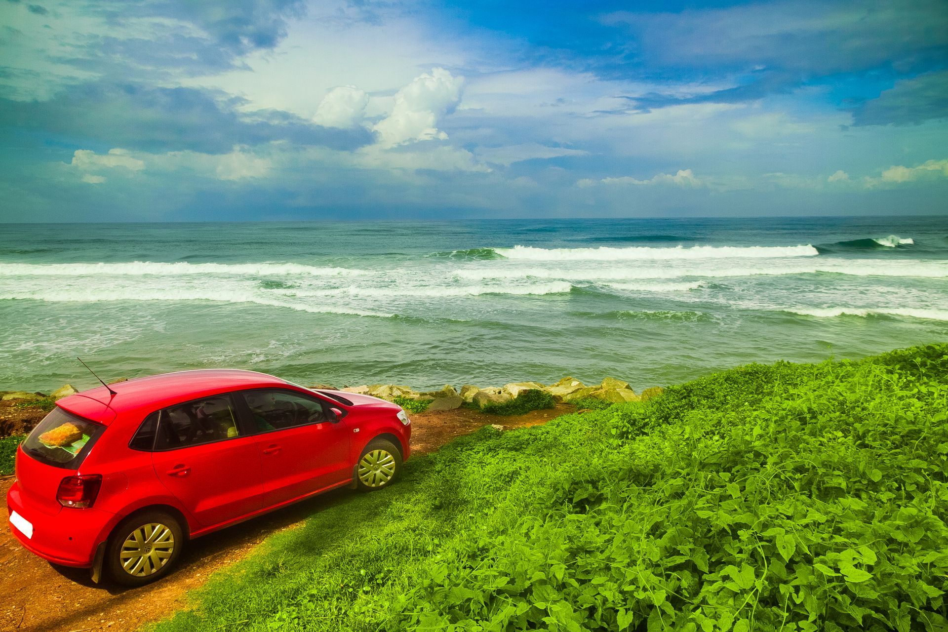 A red car is parked on a dirt road next to the ocean.