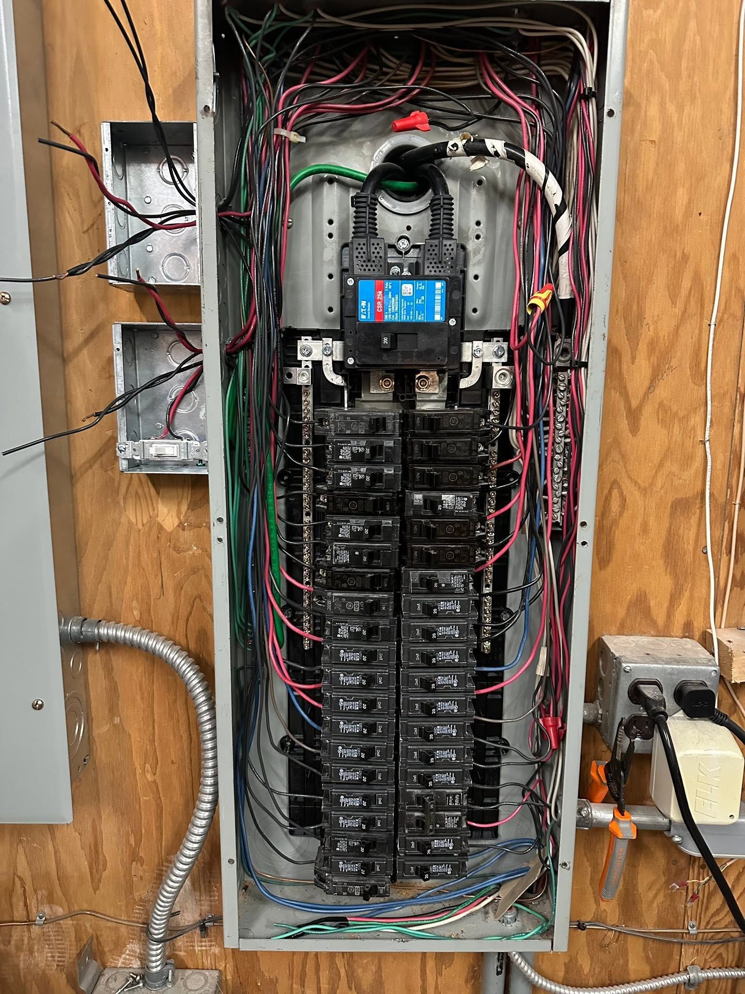 Open electrical breaker panel with many black breakers and tangled red, black, and green wires on a wooden wall.