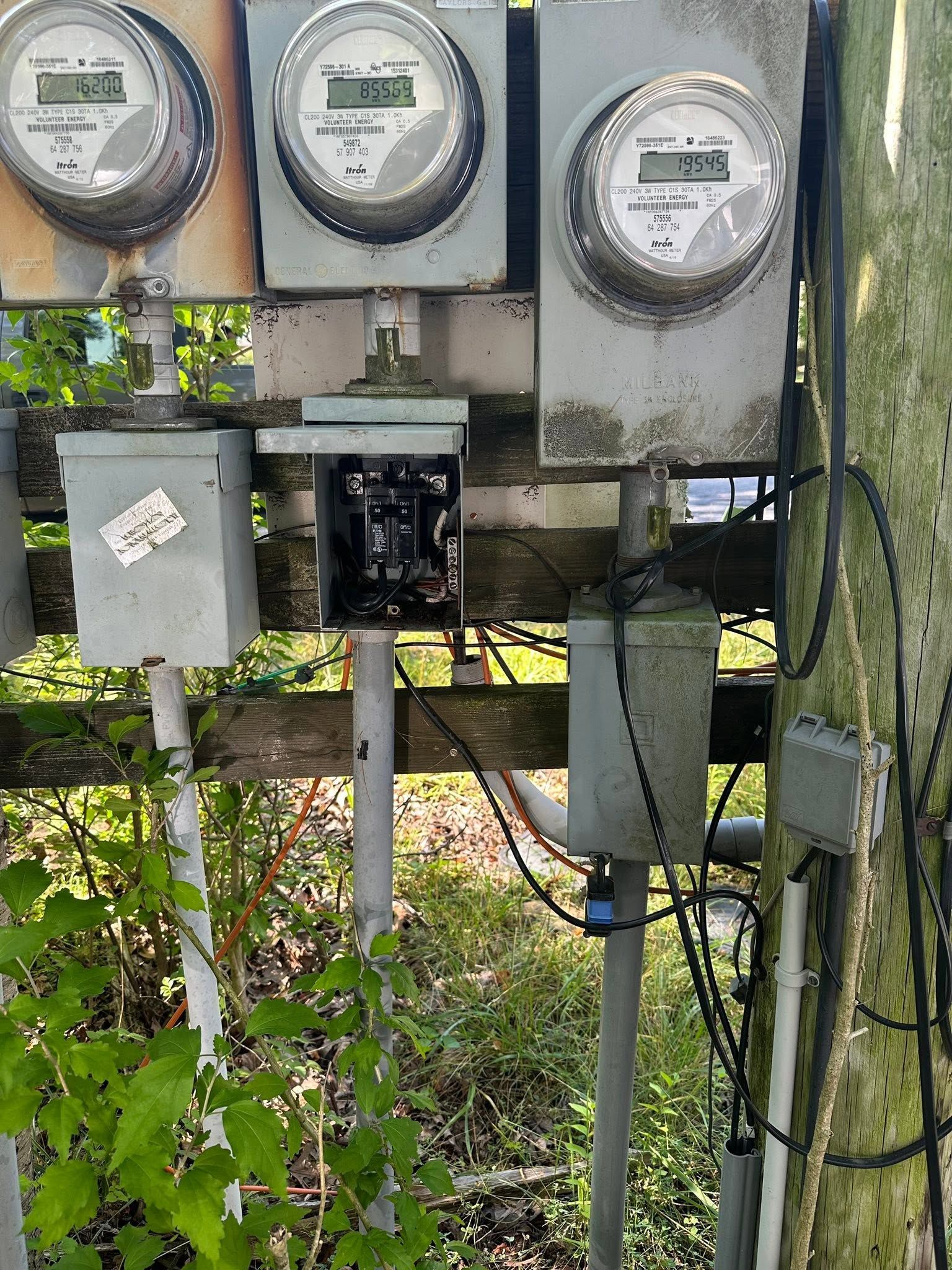 Electric meters and utility boxes mounted on a post beside a wooden fence and greenery