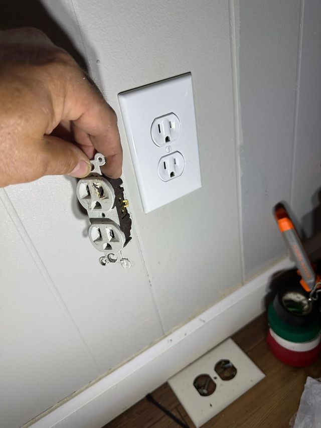 Hand removing a damaged wall outlet beside a white electrical outlet and baseboard in a room.