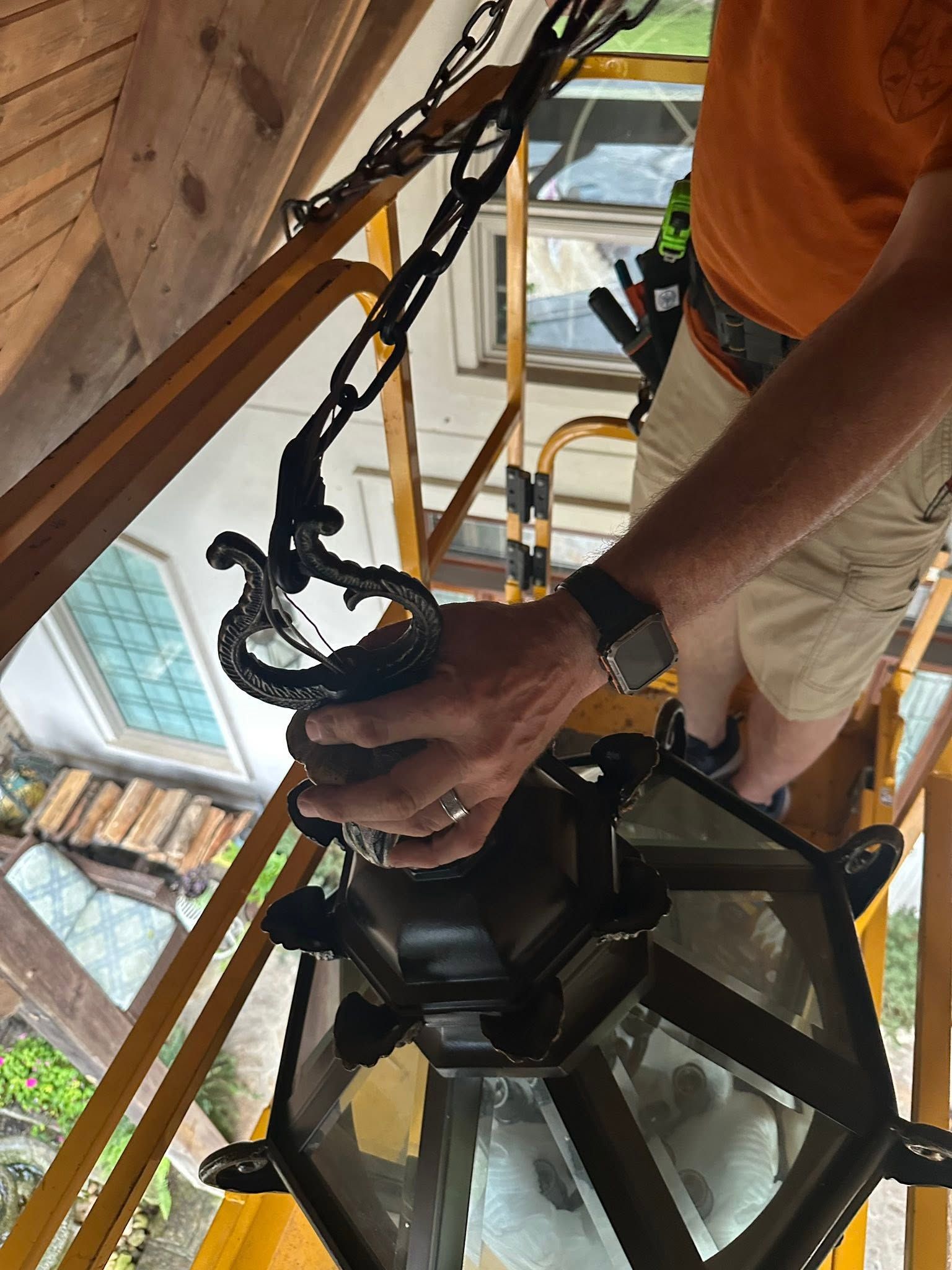 Person adjusting a hanging lantern under a wooden porch, with a chain and window visible.