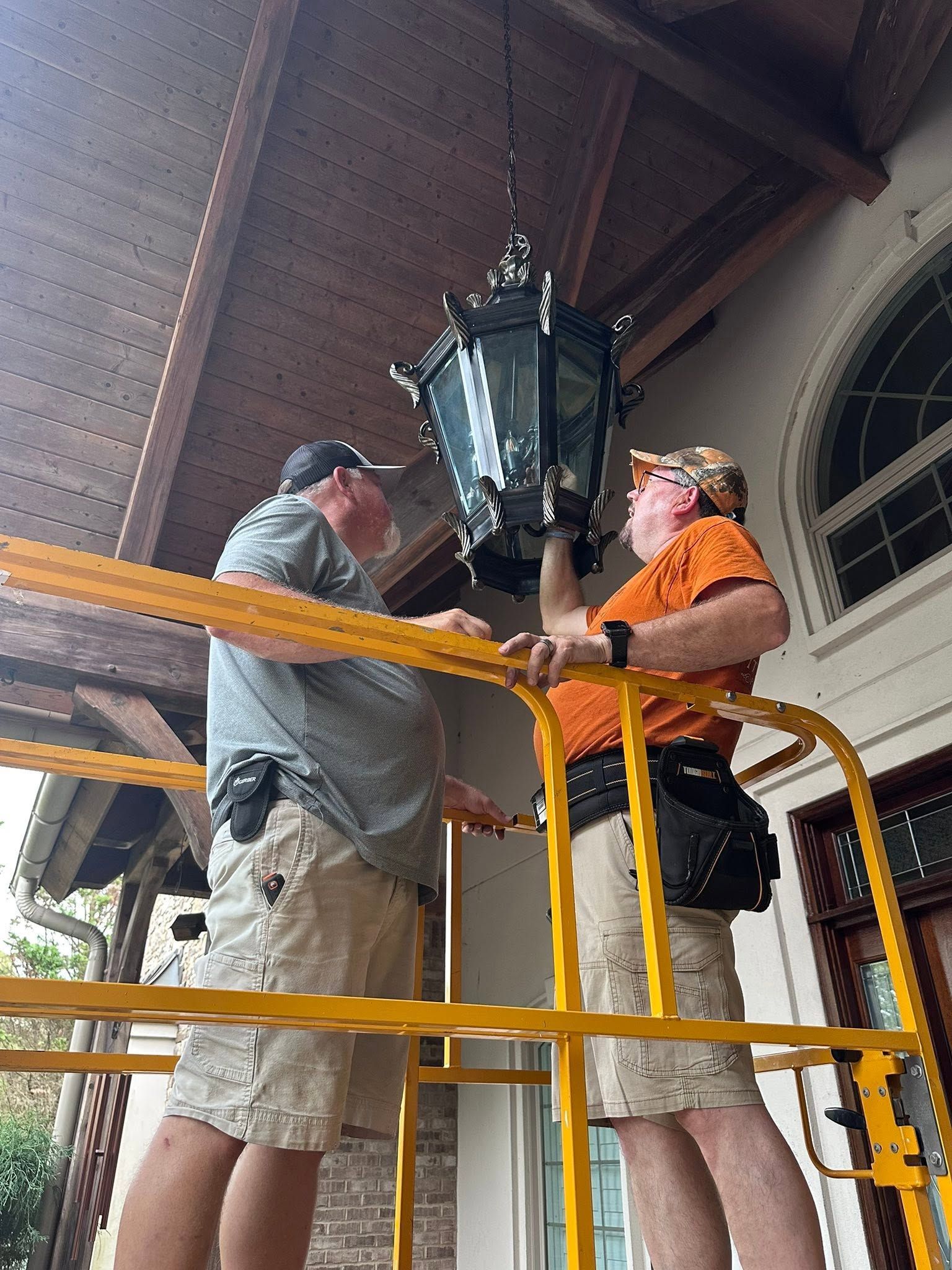 Two people replace a hanging lantern on a porch, standing on a yellow-railed lift under a wooden ceiling.