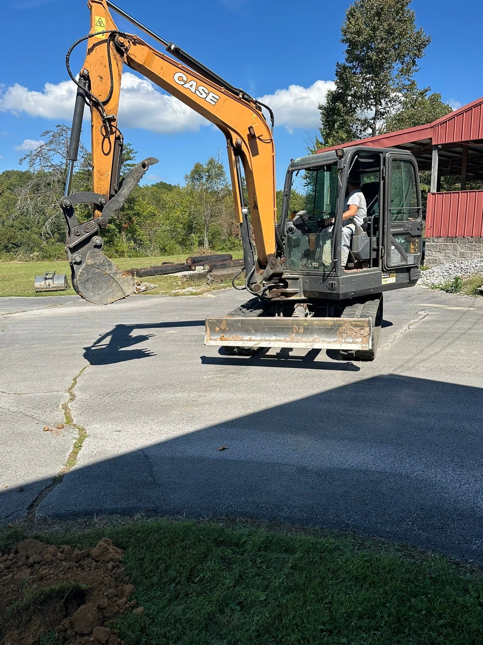 Orange excavator on a flatbed trailer beside a red building, working in a gravel lot.