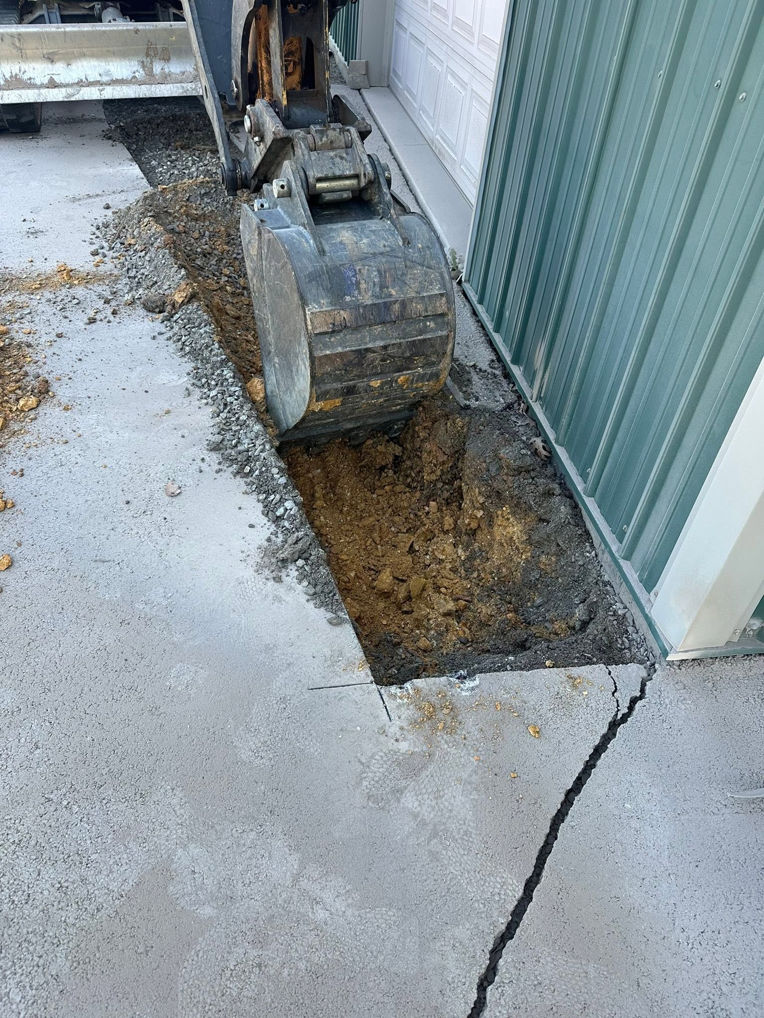 Excavator digging a trench beside a corrugated metal wall on a concrete surface
