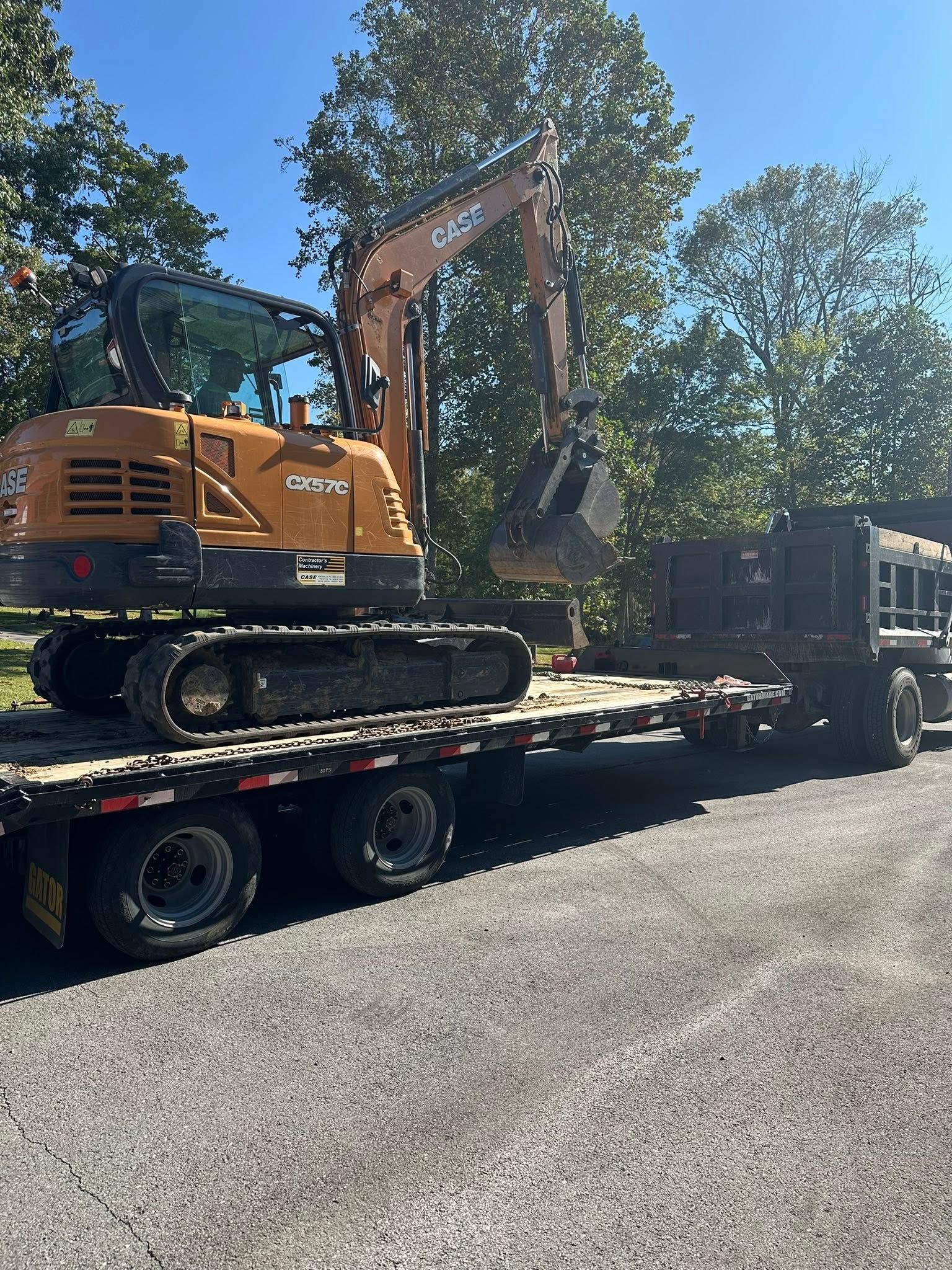 Yellow excavator loaded on a flatbed trailer beside trees under a bright blue sky.