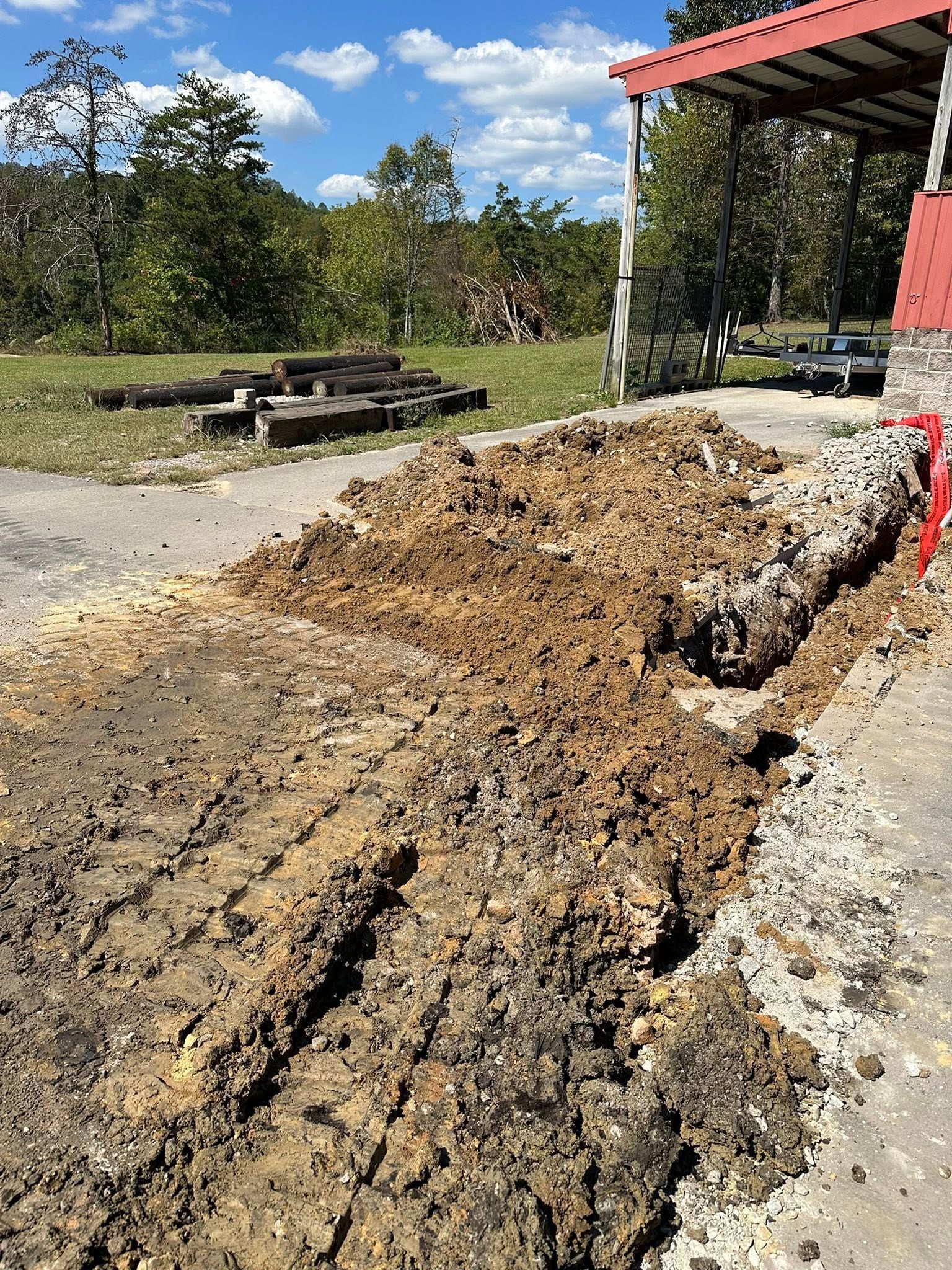 Freshly dug trench with piles of dirt beside a red-roofed outdoor shelter and picnic table