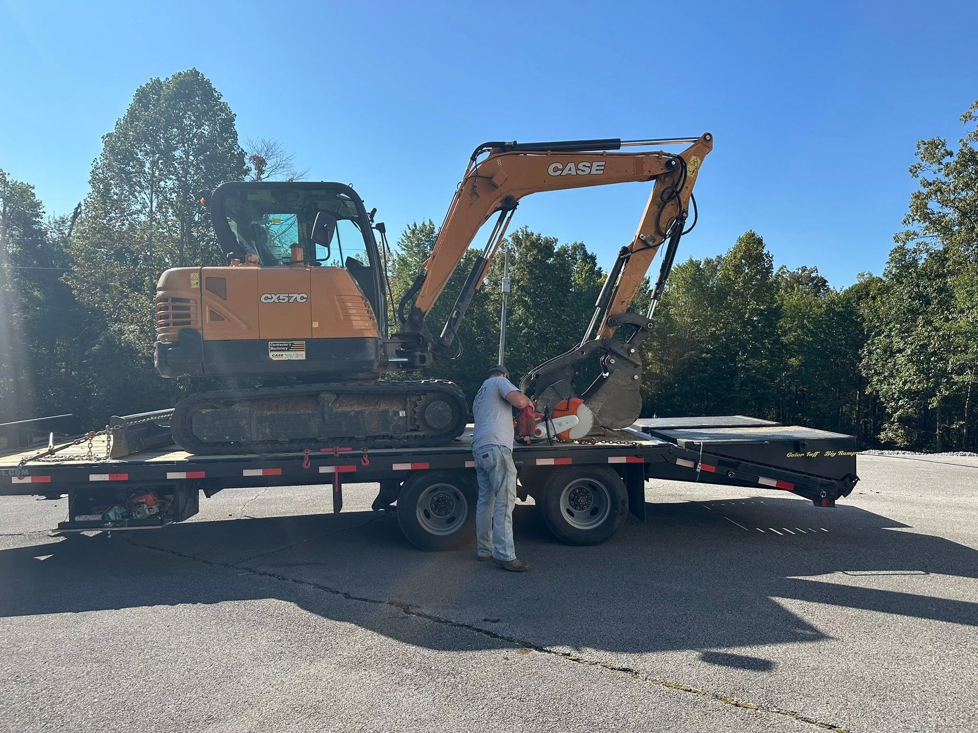 Orange excavator on a flatbed trailer with a worker in white beside it outdoors under a blue sky