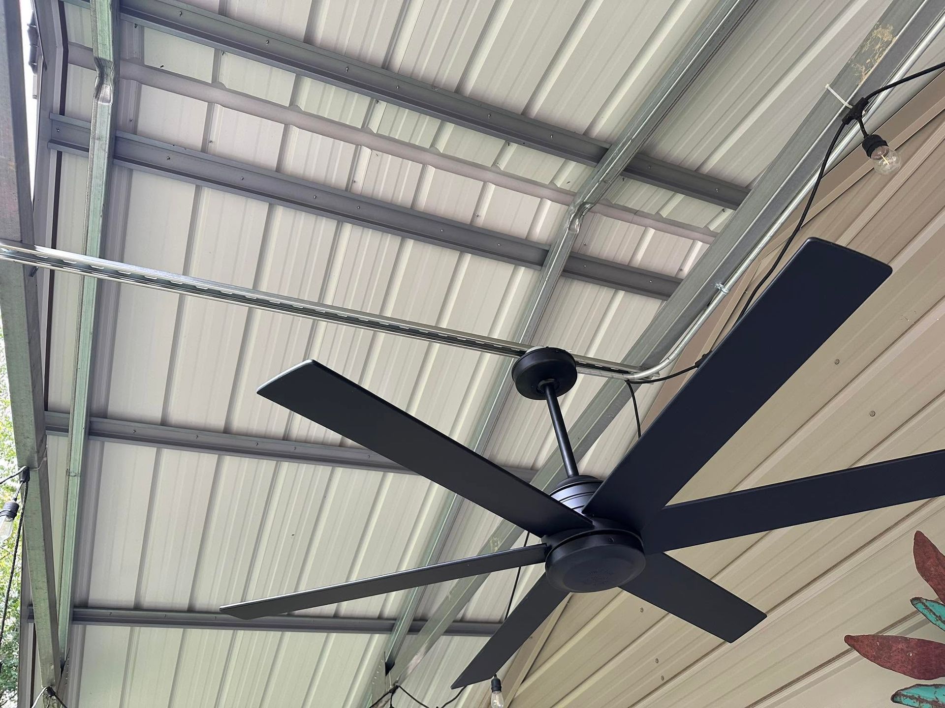 Black ceiling fan with four blades on a corrugated metal roof in a covered outdoor space