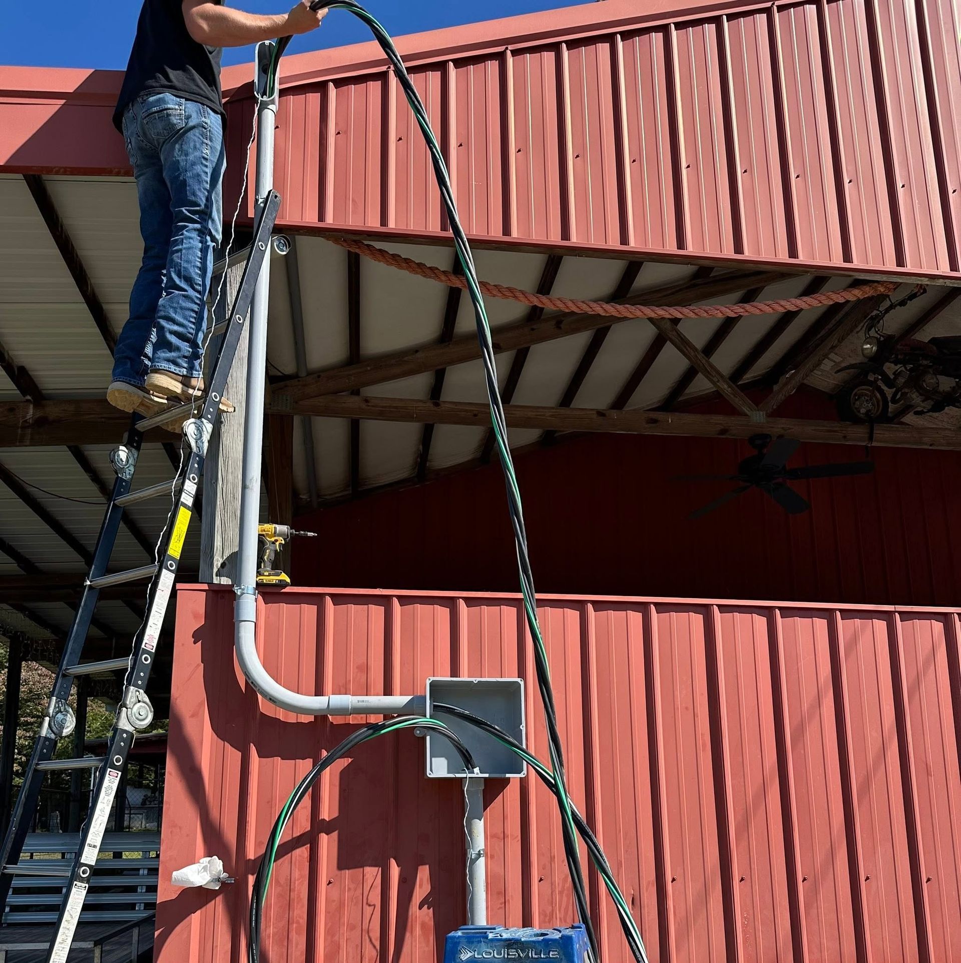 Worker on ladder inspecting red metal building exterior with exposed wiring and conduit