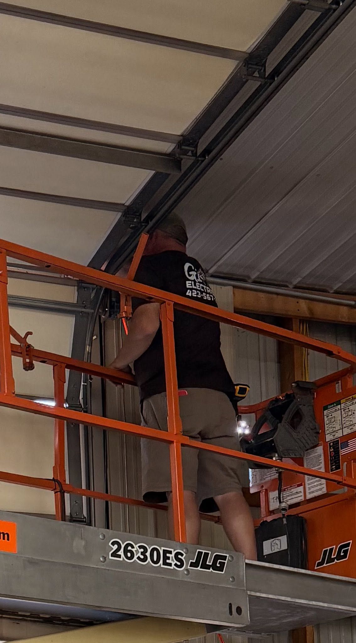 Person standing on an orange lift in a warehouse, facing shelves near the ceiling.