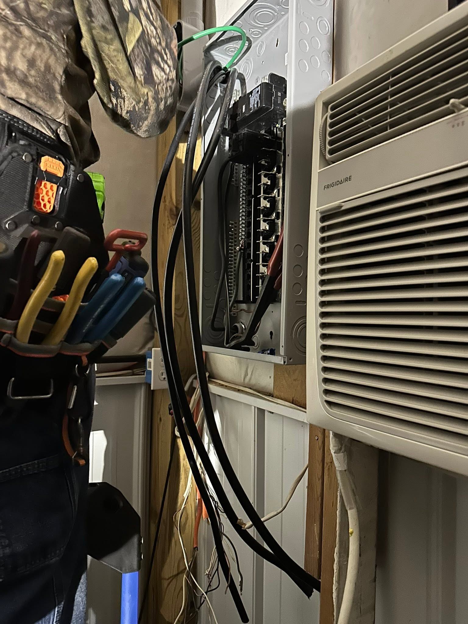 Electrical panel with exposed wires beside a wall-mounted air conditioner; tools hang on the left.