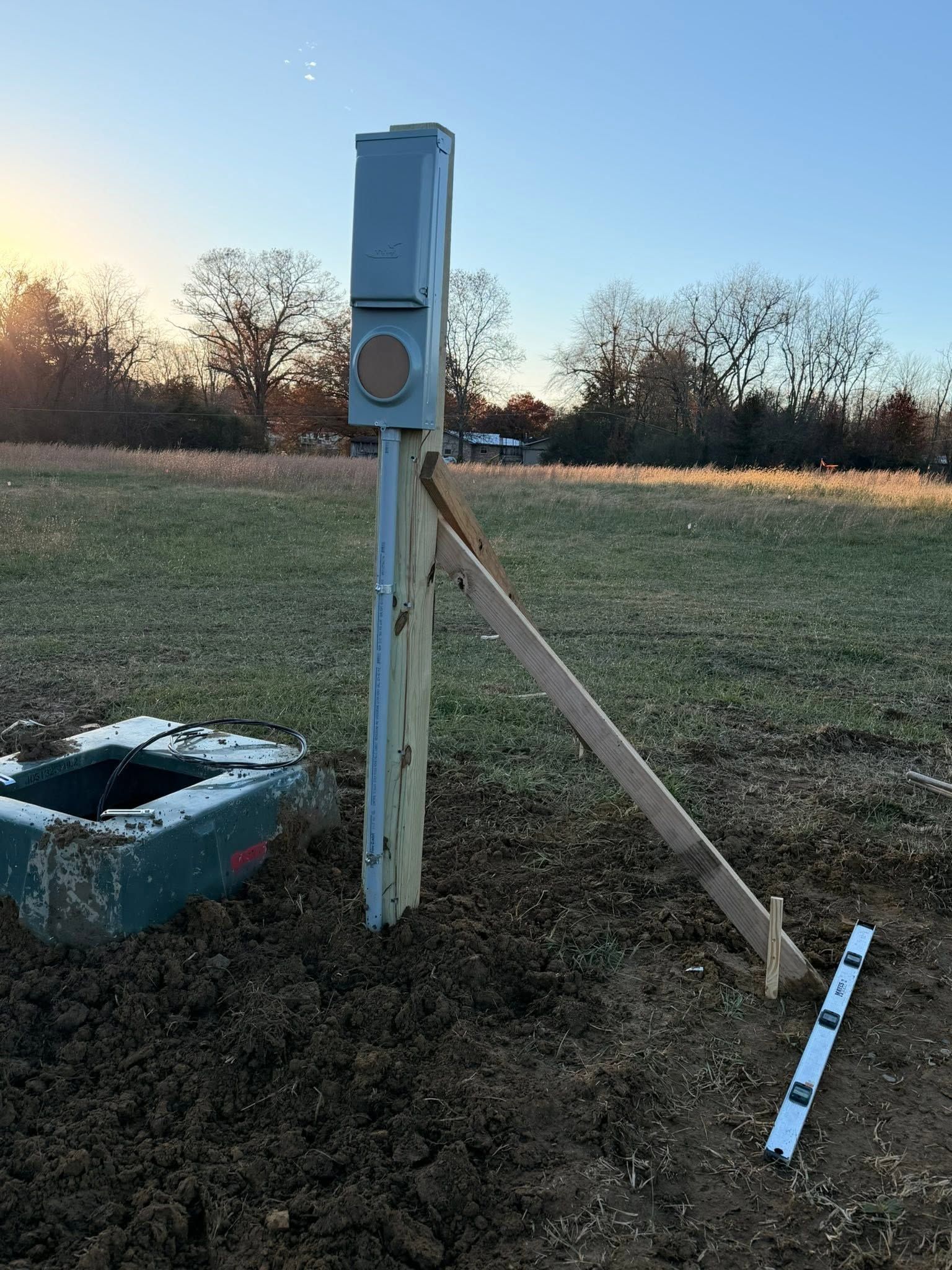 Blue utility box on a wooden post in a grassy field at sunset, with a diagonal support beam nearby