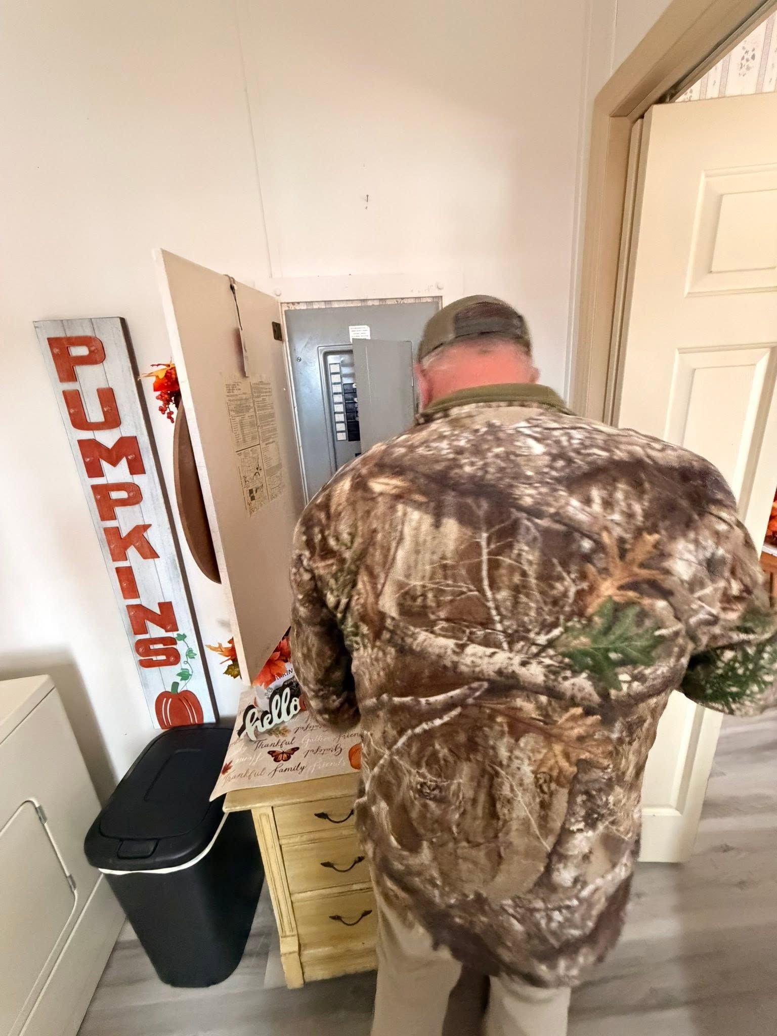 Person in camouflage jacket facing a dresser in a small room beside a door and trash bin.