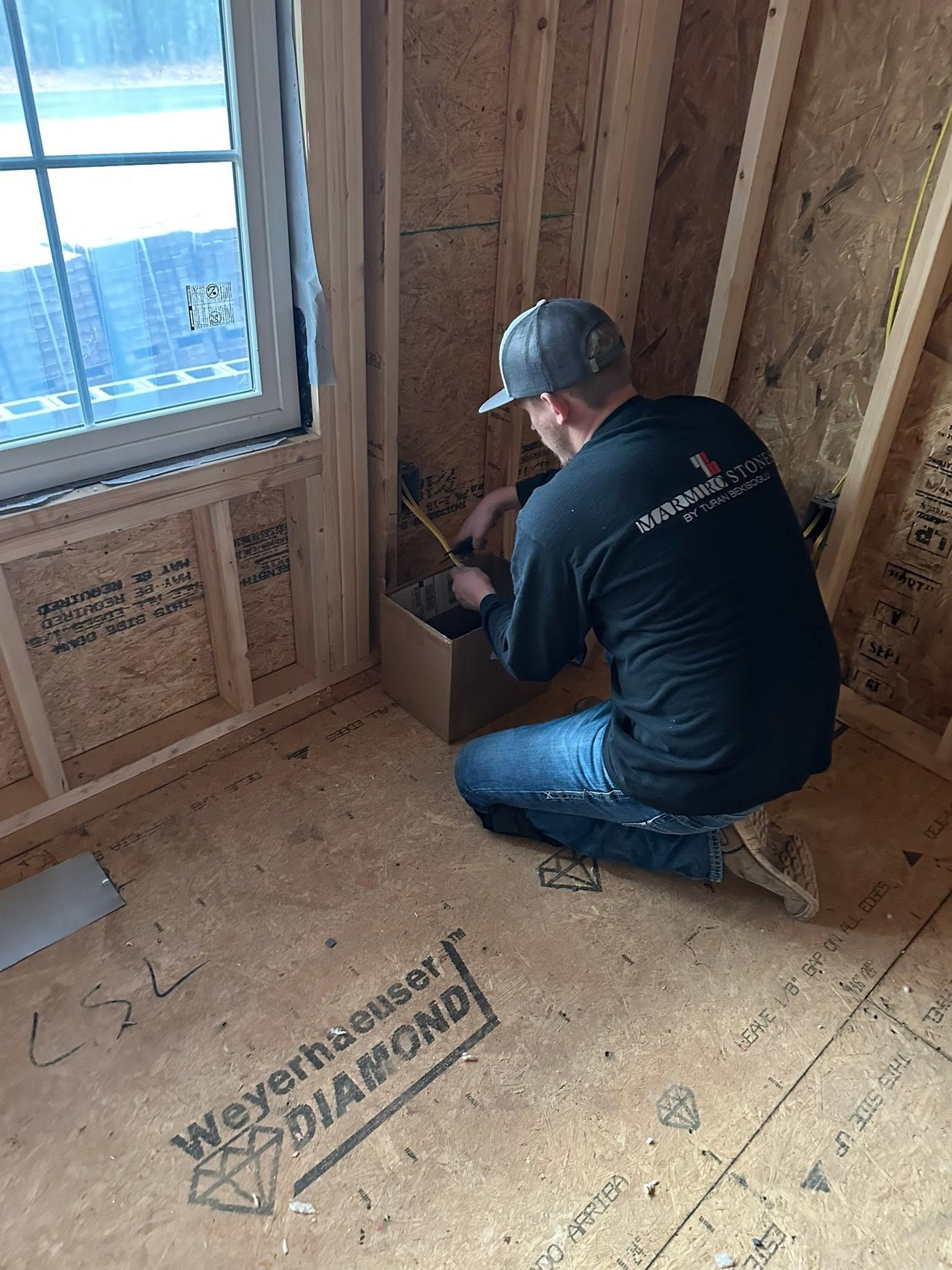 Worker kneeling in an unfinished room, installing a box near a window and exposed wood framing.
