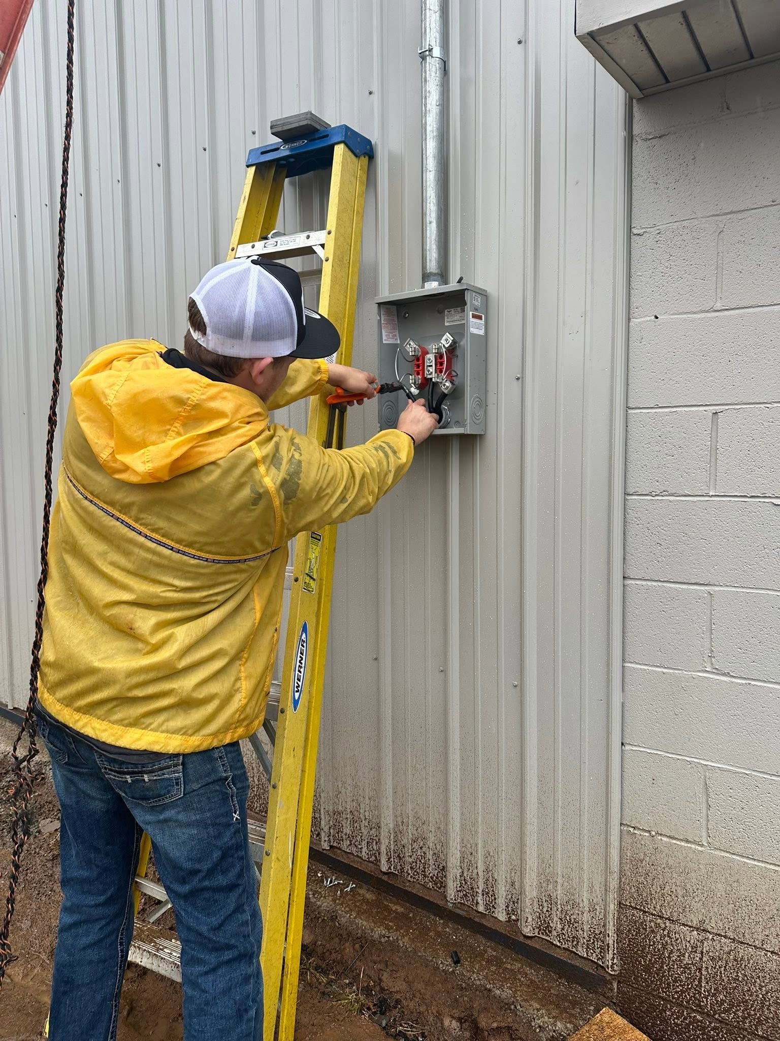 Worker in yellow rain jacket using an electrical panel on a ladder beside a corrugated metal wall.