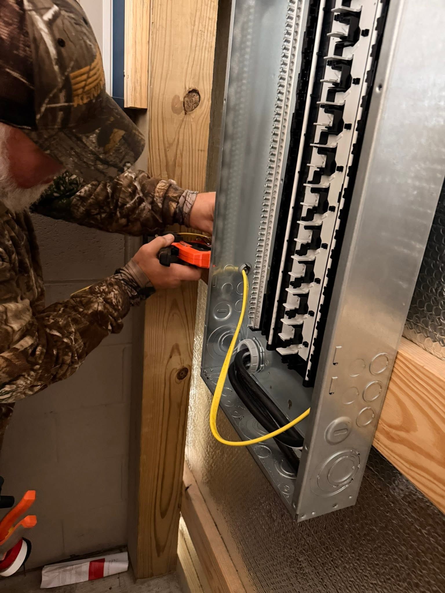 Person wiring an electrical panel inside a wall with a drill and yellow cable.