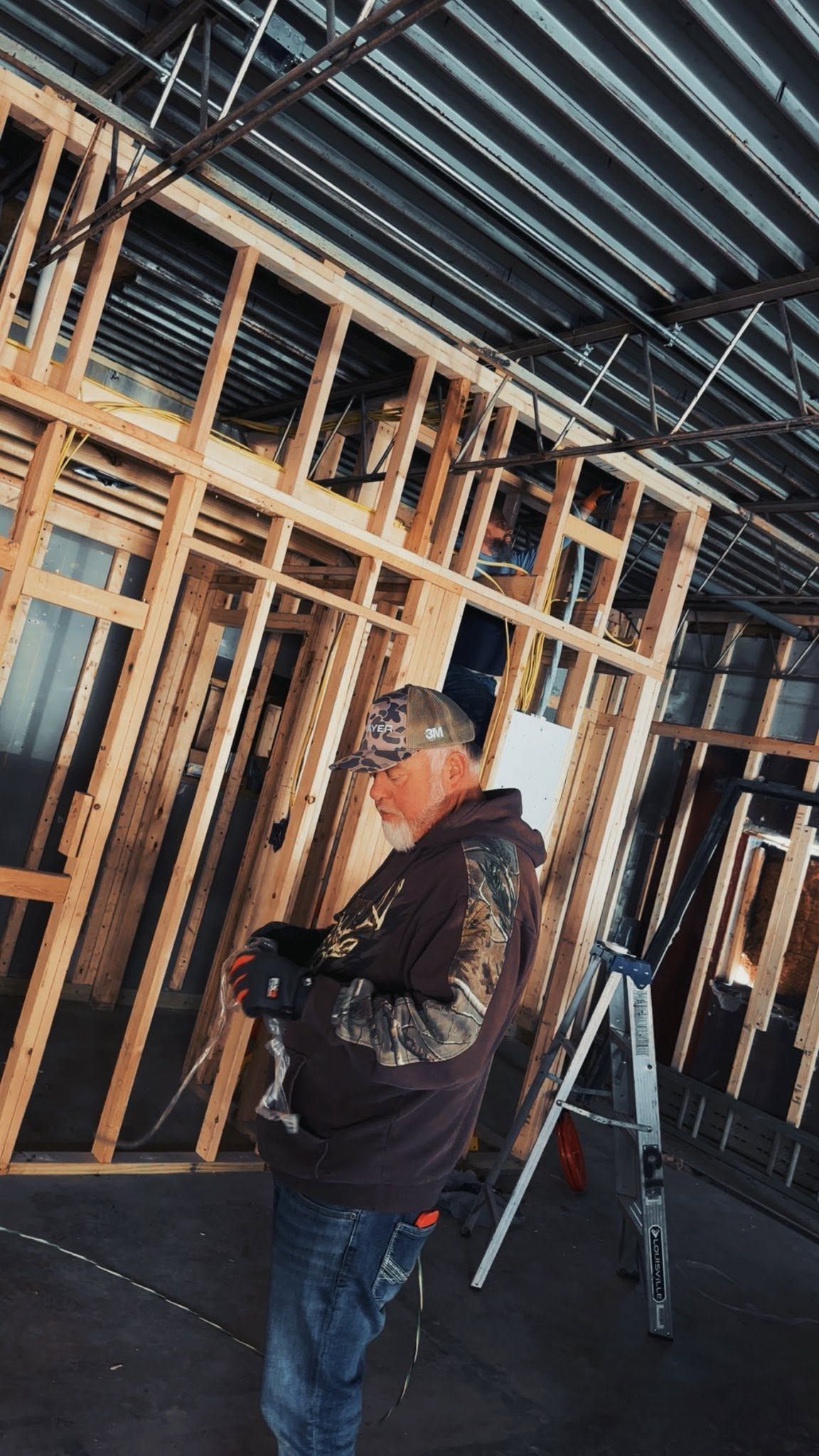 Worker in a hard hat using a handheld device inside a framed construction site with exposed studs and a ladder