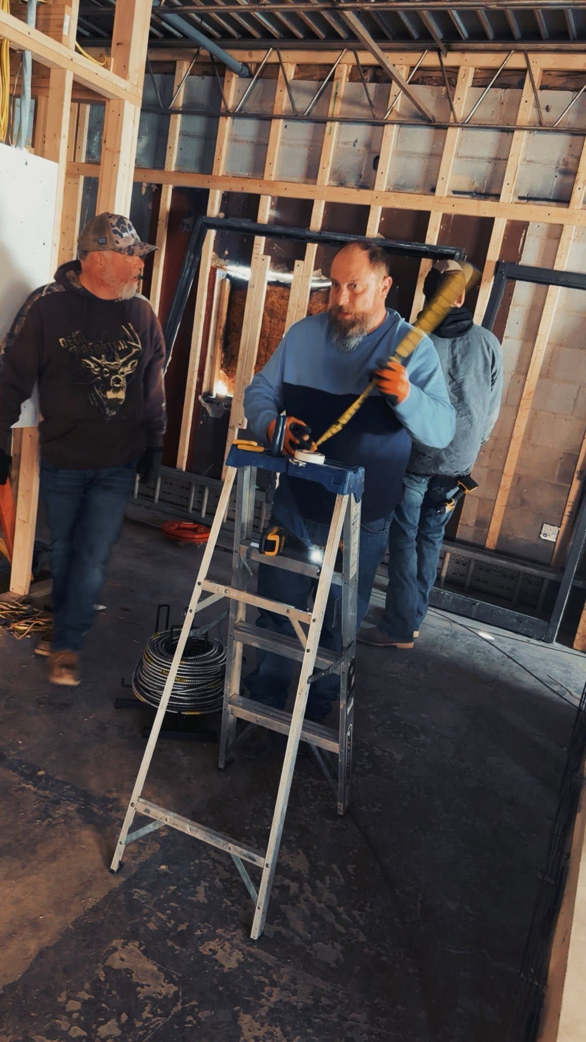 Three workers on ladders inside a room with exposed wall framing, handling electrical wiring.