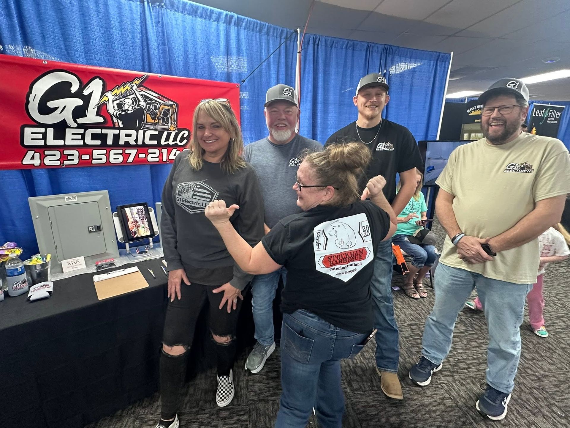 People at a trade booth with a G1 Electric sign; one woman poses while another kneels beside her.