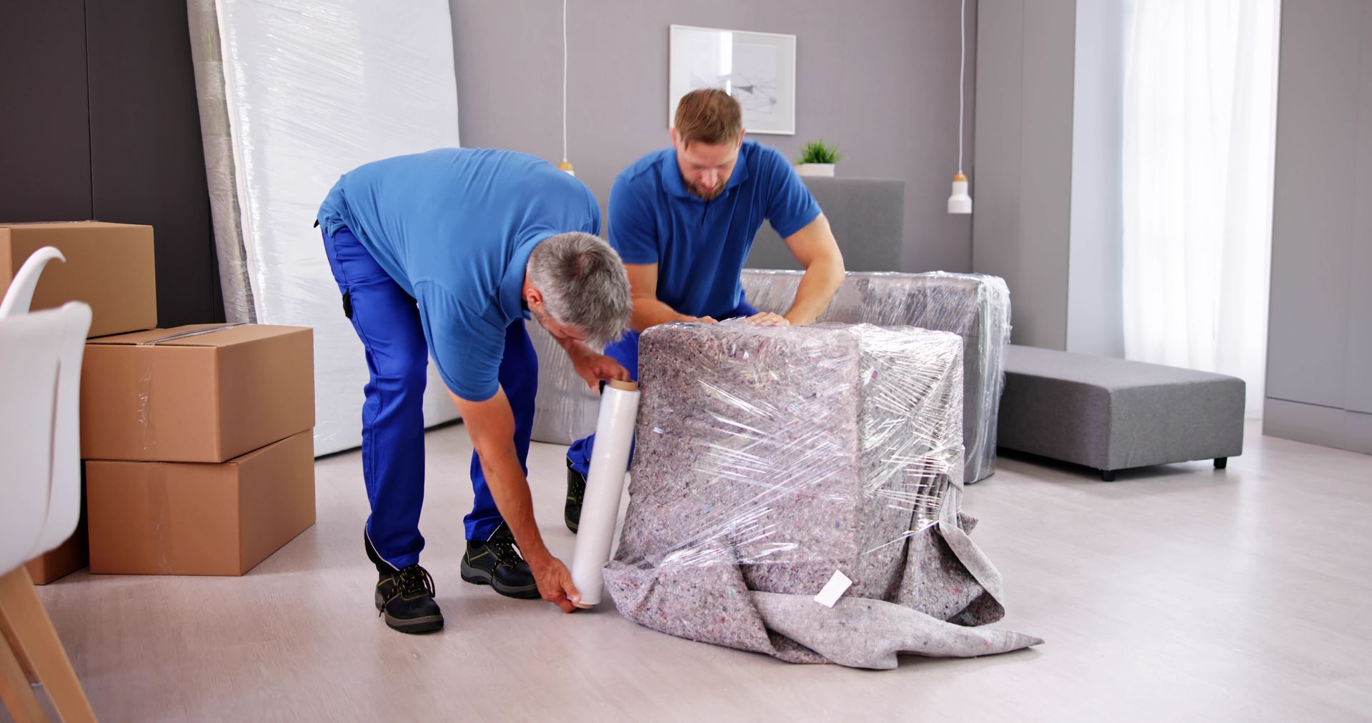 Two movers in blue uniforms wrap a piece of furniture in plastic film inside a room with cardboard boxes.