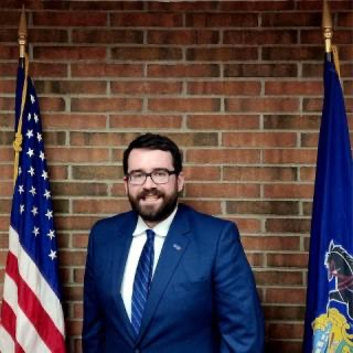 Man in blue suit, smiling, standing between US and state flags, brick wall background.