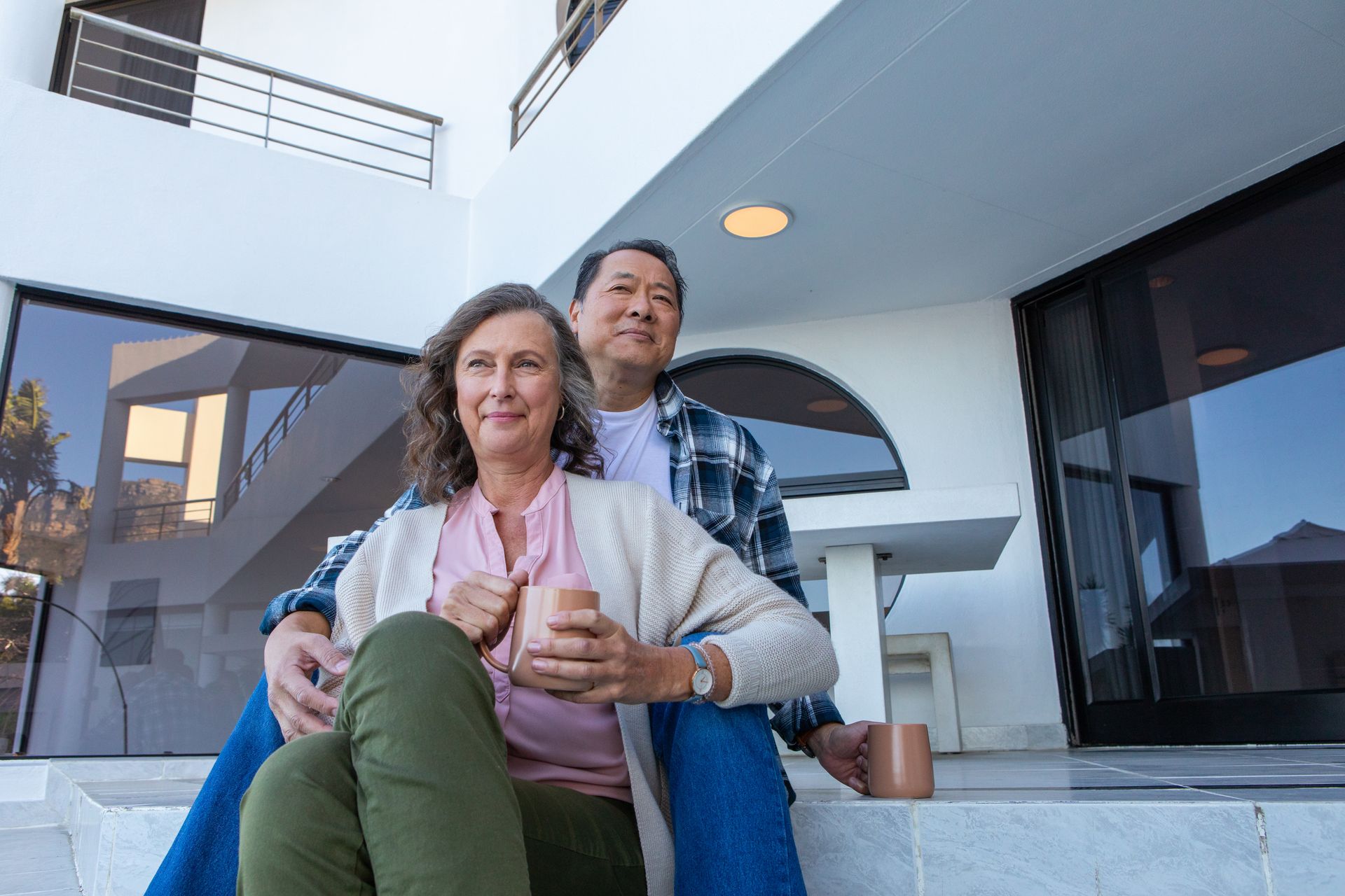 A couple is sitting on steps outside their home holding mugs. The home’s windows are tinted.