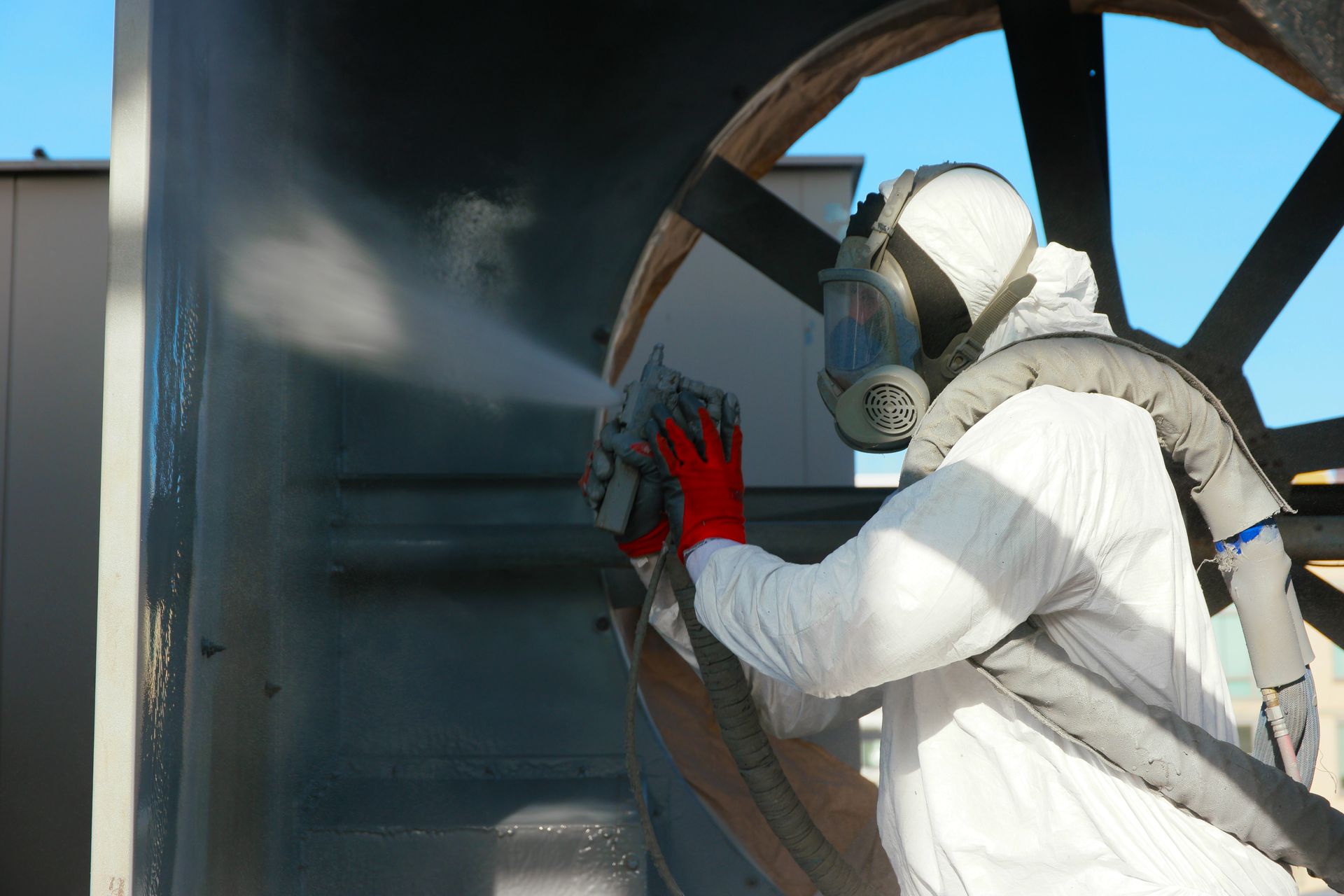 Worker in protective gear applying industrial coating with spray equipment to a large metal surface.