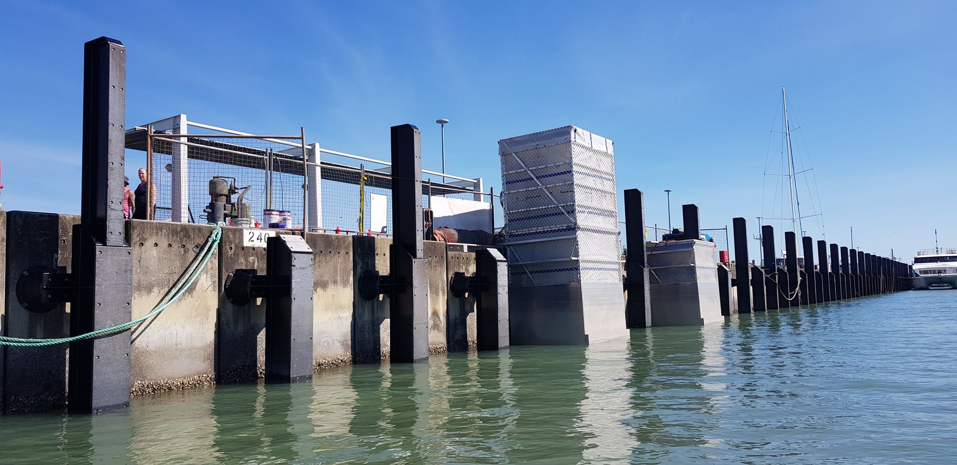 Dock pilings and a metal walkway at a harbor on calm water under a clear blue sky