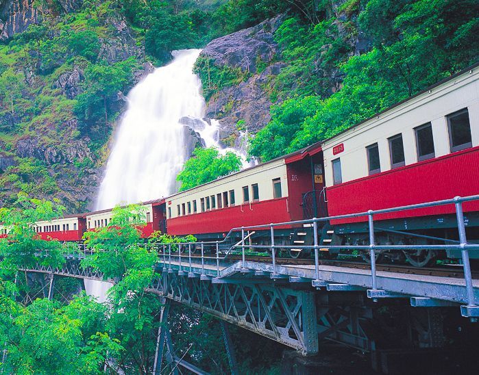 Red-and-cream train crossing a bridge beside a tall waterfall in a lush green mountainside