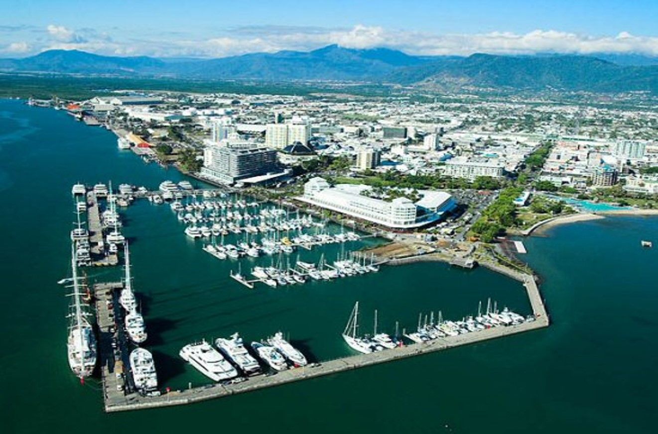 Aerial view of a marina with boats, city buildings, and mountains along a blue coastline.