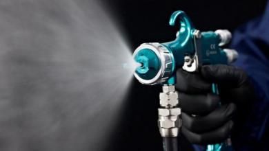 Black-gloved hand sprays mist from a teal spray gun against a dark background