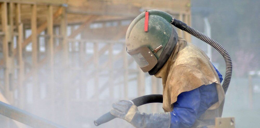 Worker in protective gear sandblasting metal in a dusty workshop, wearing a face shield and gloves