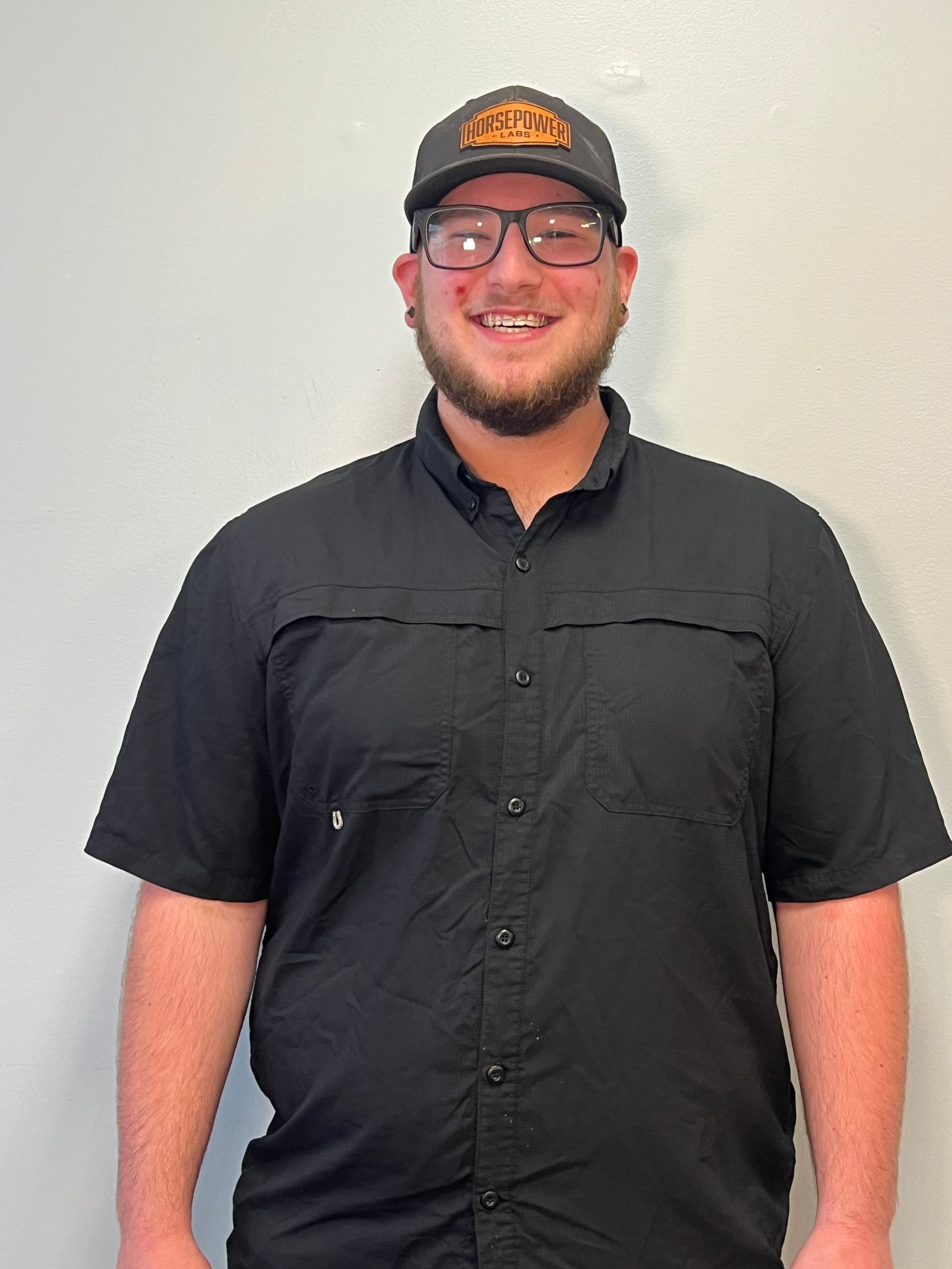 Man wearing a black shirt and cap, smiling and standing against a plain light wall.