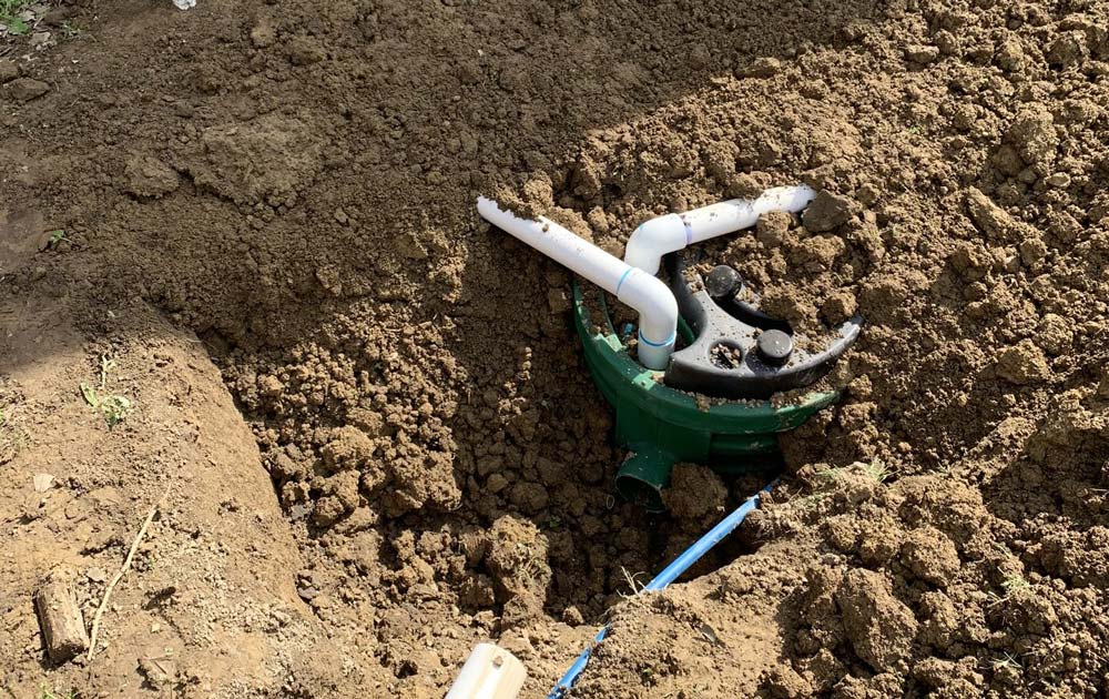 Green sprinkler valve in a dirt trench with white PVC pipes attached